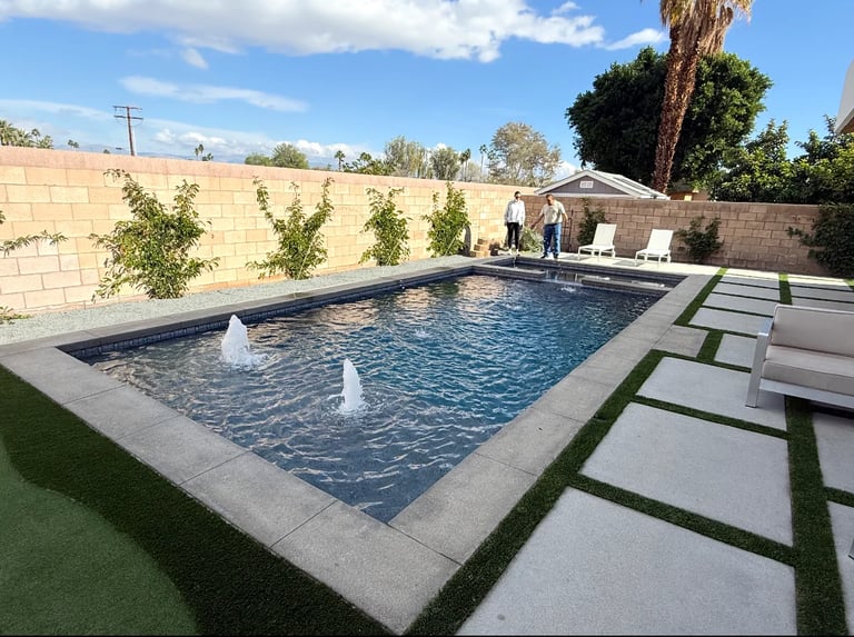 Backyard swimming pool with water fountains, concrete deck, and artificial turf surrounded by stone walls and landscaping
