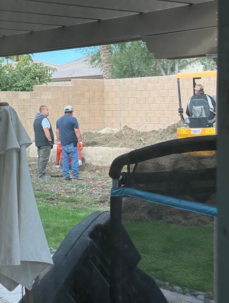 Construction workers and small excavator at a residential backyard job site behind a tan brick wall