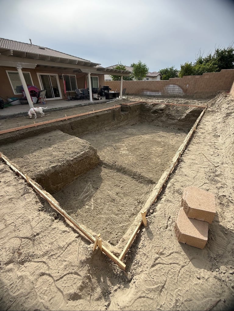 Excavated foundation pit in sandy backyard with wooden framing and single-story house with patio in background