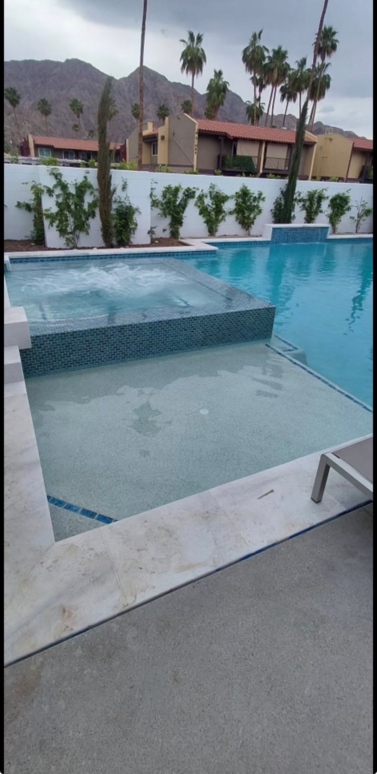 Modern resort pool with shallow wading section and deep end, surrounded by palm trees and desert mountains under cloudy sky