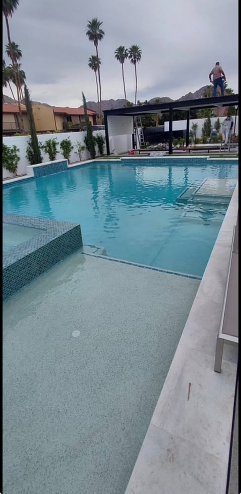 Modern residential swimming pool with turquoise water, concrete deck, palm trees, and mountains in the background under cloudy sky