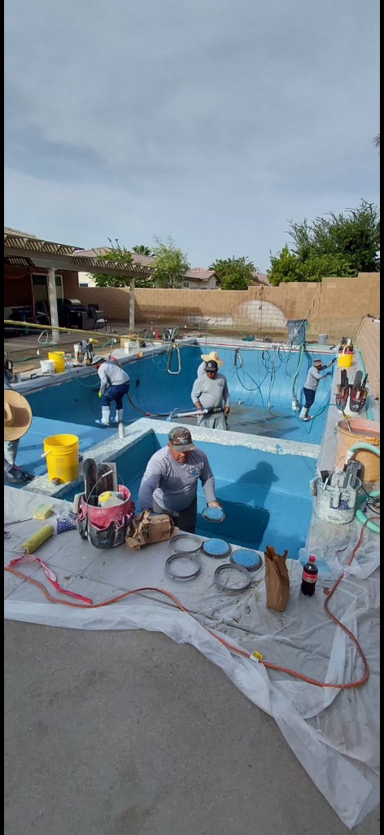Workers resurfacing an empty residential swimming pool with blue coating, surrounded by equipment and tools